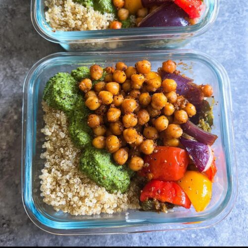 Close-up of a glass container showing a Chickpea Lunch Meal Prep with seasoned chickpeas, quinoa, green sauce, and roasted vegetables.