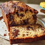 A close-up of a freshly baked Chocolate Chunk Banana Bread loaf, with one slice cut and resting beside it on a plate.