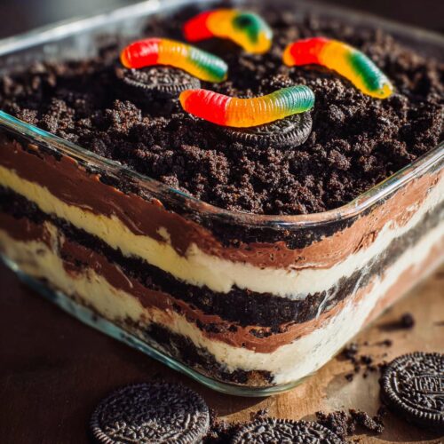 Close-up of a glass dish filled with layered Classic Dirt Cake, topped with crushed cookies and gummy worms.