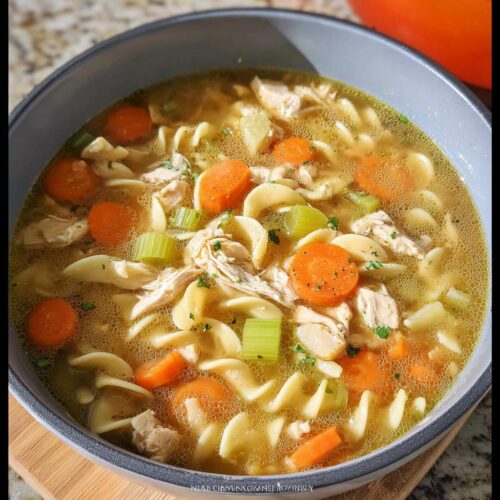 A close-up of a bowl filled with hot Cozy Turkey & Chicken Soup featuring shredded meat, egg noodles, carrots, and celery.