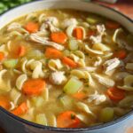 Close-up of steaming Cozy Turkey & Chicken Soup filled with egg noodles, sliced carrots, celery, and shredded poultry in a blue enameled pot.