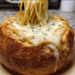 A close-up of a Creamy Alfredo Spaghetti Bread Bowl topped with melted mozzarella and parsley, with spaghetti being lifted by a fork.