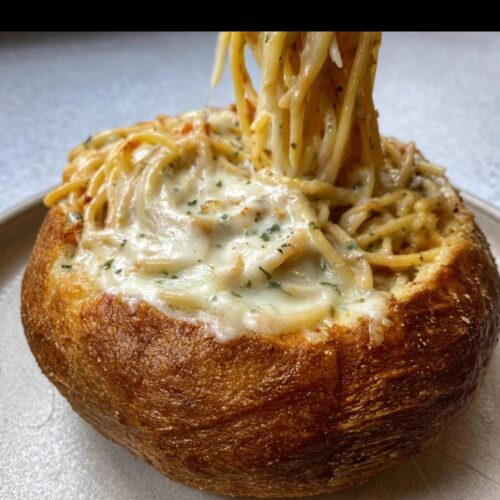 A close-up of a Creamy Alfredo Spaghetti Bread Bowl with spaghetti being lifted out, showing melted cheese.