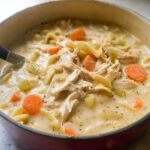 Close-up of rich Creamy Chicken Noodle Soup for Dinner featuring shredded chicken, egg noodles, carrots, and celery in a pot.