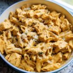 Close-up of creamy chicken pasta meal prep featuring penne pasta, seasoned chicken chunks, and melted cheese in a light blue serving dish.