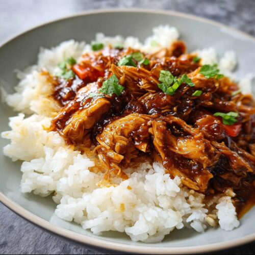 A bowl of fluffy white rice topped with shredded Crockpot Teriyaki Chicken coated in a rich, dark sauce and garnished with green herbs.