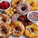 A platter featuring various donuts, bacon, raspberries, and bowls of sprinkles, perfect for a Fun Weekend Breakfast Idea.