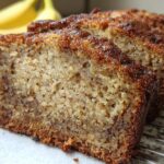 Close-up view showing the texture of slices of Extra Moist Banana Bread cooling on a wire rack, with bananas blurred in the background.