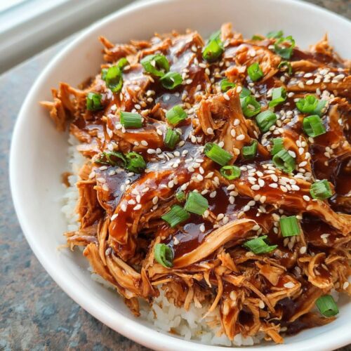 A white bowl filled with rice topped with shredded Family-Friendly Crockpot Teriyaki Chicken, garnished with sesame seeds and green onions.