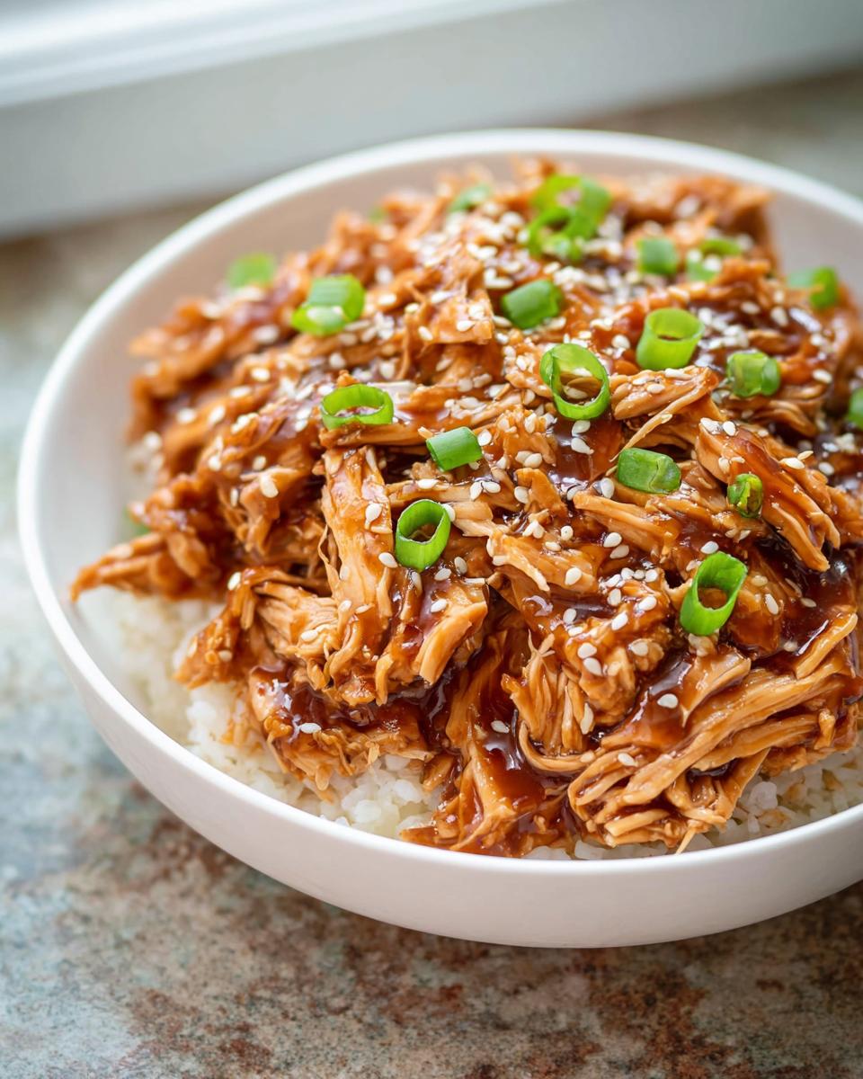 A bowl of shredded Family-Friendly Crockpot Teriyaki Chicken glazed with sauce, served over white rice and topped with sesame seeds and green onions.