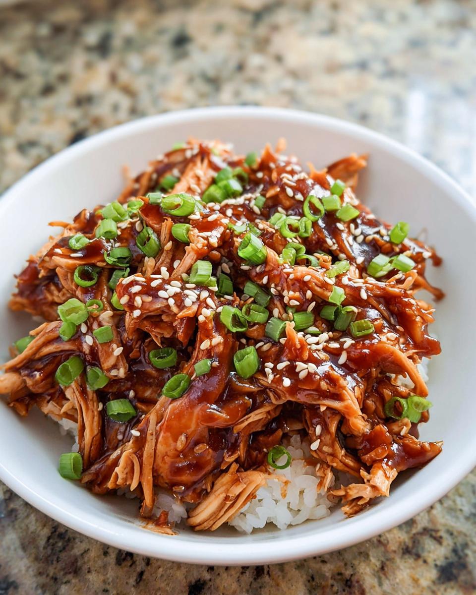A bowl of shredded Family-Friendly Crockpot Teriyaki Chicken served over white rice, topped with sesame seeds and green onions.
