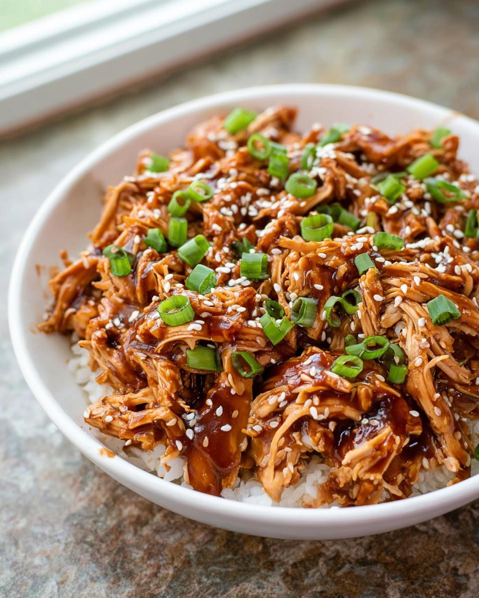A white bowl filled with shredded Family-Friendly Crockpot Teriyaki Chicken served over rice, topped with sesame seeds and green onions.