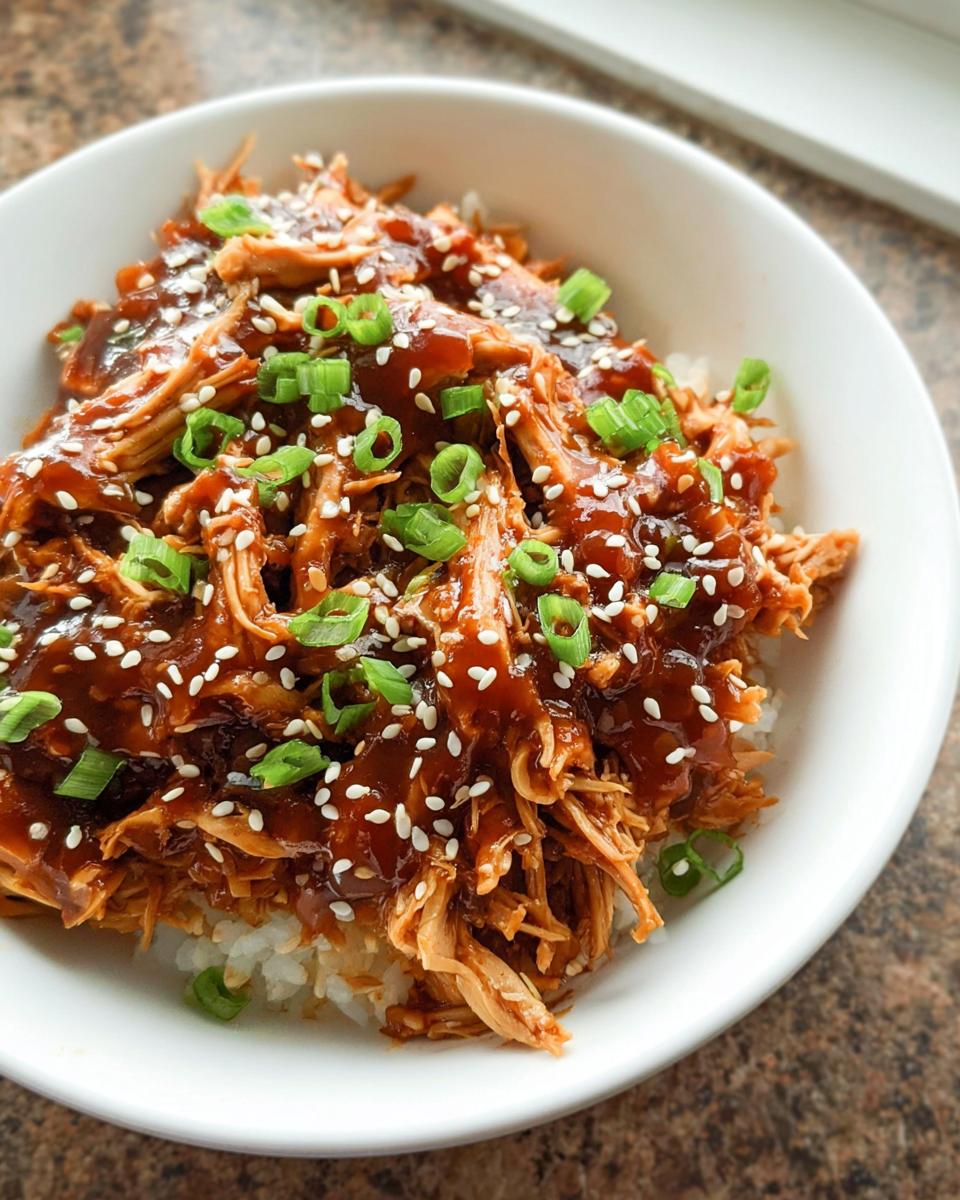 Close-up of shredded Family-Friendly Crockpot Teriyaki Chicken served over white rice, topped with teriyaki glaze, sesame seeds, and green onions.