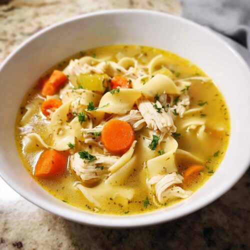 Close-up of a white bowl filled with rich broth, egg noodles, shredded chicken, and bright orange carrots in Family-Style Chicken Noodle Soup.