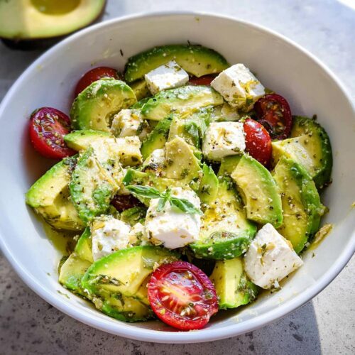 A vibrant bowl filled with sliced avocado, cubes of feta cheese, and halved cherry tomatoes, dressed for a Feta & Avocado Lunch Bowl.