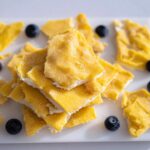 A stack of bright yellow Frozen Lemon Yogurt Bark pieces garnished with fresh blueberries on a white platter.