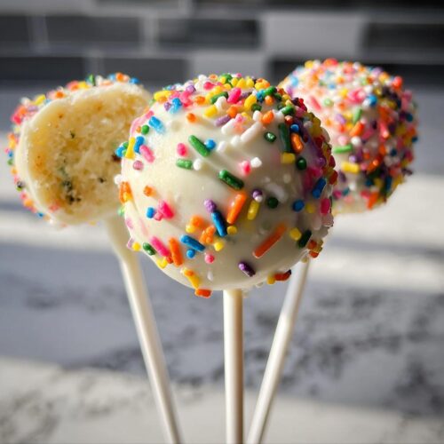 Three Funfetti Cake Pops dipped in white coating and covered in colorful sprinkles, one is cut open.