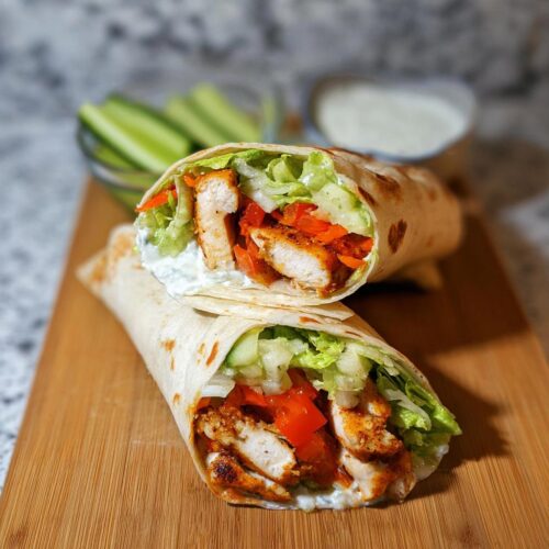 Two halves of a grilled chicken wraps filled with seasoned chicken, lettuce, tomatoes, and creamy sauce, resting on a wooden board.