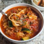 A close-up view of a white bowl filled with rich, tomato-based Healthy Lasagna Soup, featuring ground meat, zucchini chunks, and wide noodles.