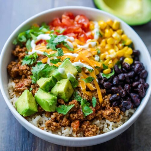A vibrant bowl featuring brown rice topped with seasoned ground meat, black beans, corn, avocado, cheese, and sour cream for a Healthy Taco Rice Bowl.
