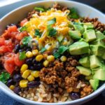 A close-up of a Healthy Taco Rice Bowl featuring seasoned ground meat, brown rice, black beans, corn, diced tomatoes, avocado, and shredded cheese.