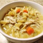 A close-up view of a bowl filled with Hearty Creamy Chicken Soup featuring egg noodles, shredded chicken, carrots, and celery.