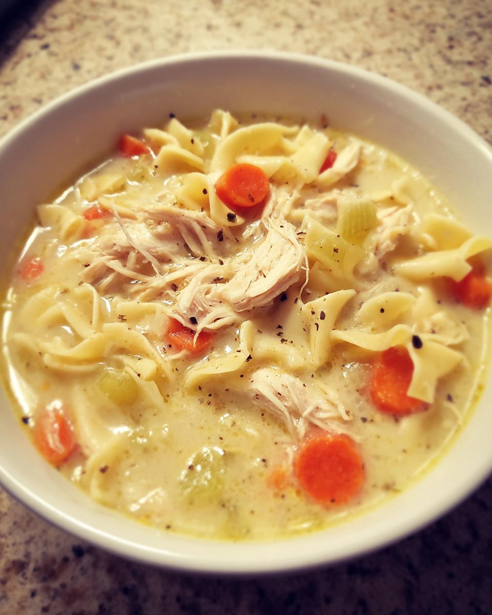 Close-up of a white bowl filled with Hearty Creamy Chicken Soup featuring shredded chicken, egg noodles, carrots, and celery.