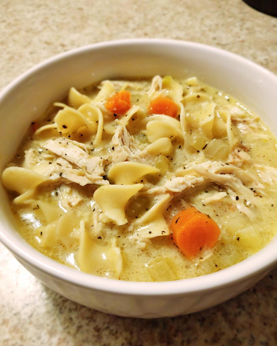 A close-up view of a bowl filled with Hearty Creamy Chicken Soup featuring egg noodles, shredded chicken, carrots, and celery.