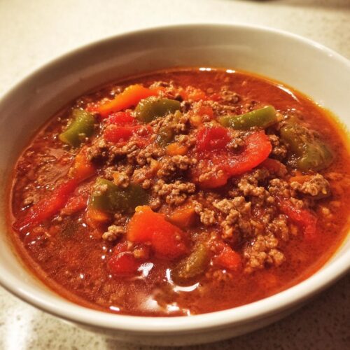 Close-up of a white bowl filled with Hearty Stuffed Pepper Soup, featuring ground meat and chunks of red and green peppers in a rich tomato broth.