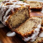 Close-up of a sliced Iced Cinnamon Banana Loaf drizzled with thick white glaze on a wooden board.
