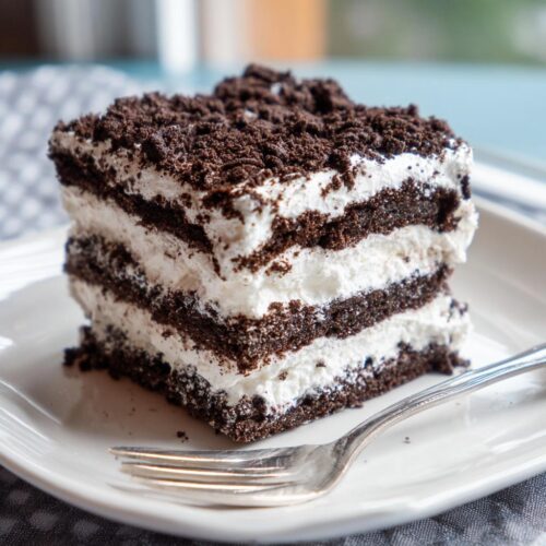A square slice of No-Bake Oreo Icebox Cake showing layers of dark chocolate wafers and white cream, topped with crushed Oreos.