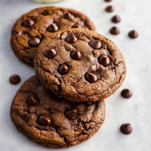 Three rich, fudgy One-Bowl Chocolate Cookies stacked on a white surface with scattered chocolate chips.