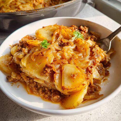 A close-up of a serving of One-Pan Hobo Casserole featuring layered potatoes, ground meat, and caramelized onions in a white bowl.