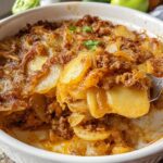 A spoonful of golden One-Pan Hobo Casserole featuring layered potatoes and ground meat being lifted from a white baking dish.