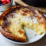 A golden, puffy Oven Pancake Breakfast dusted with powdered sugar, served on a white plate with berries nearby.