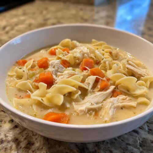 A close-up of a white bowl filled with Quick Creamy Chicken Noodle Soup, featuring egg noodles, shredded chicken, and bright orange carrots.