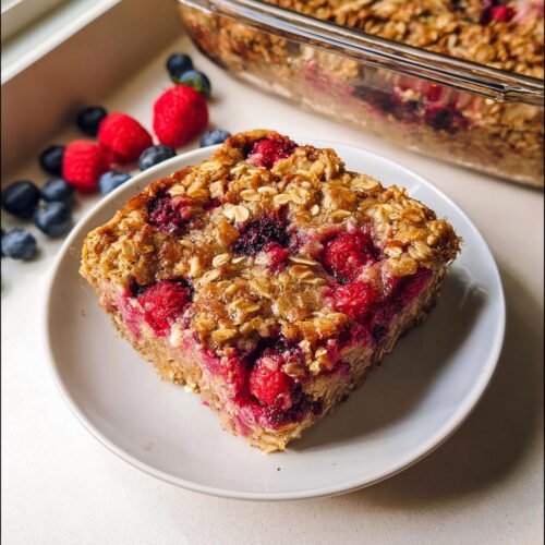A square slice of raspberry Baked Oatmeal You Can Prep Ahead on a white plate, with the baking dish visible behind it.