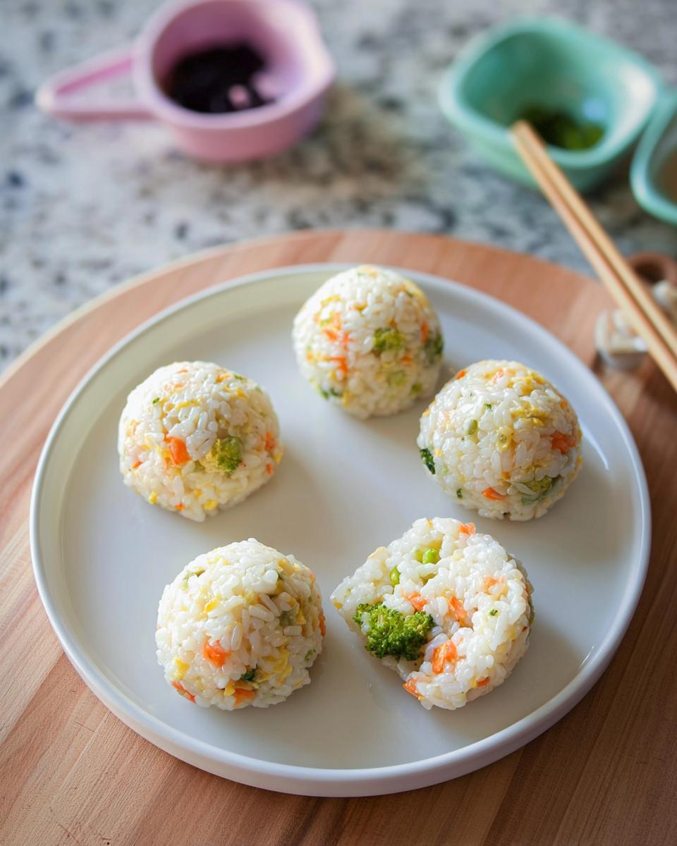 Five colorful vegetable rice ball sushi lunch portions served on a white plate, one is cut open showing broccoli and carrots.