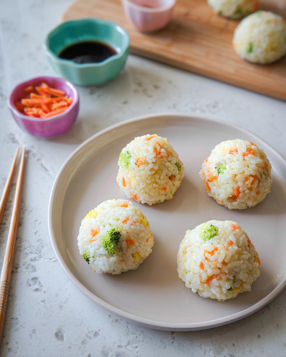 Four perfectly formed Rice Ball Sushi Lunch portions mixed with visible pieces of broccoli and shredded carrots on a light plate.