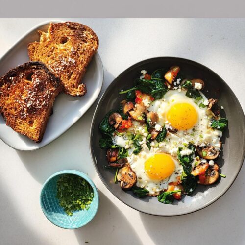 A plate featuring two sunny-side-up eggs over sautéed spinach, mushrooms, and feta, served alongside burnt toast. A perfect Savory Breakfast Idea.