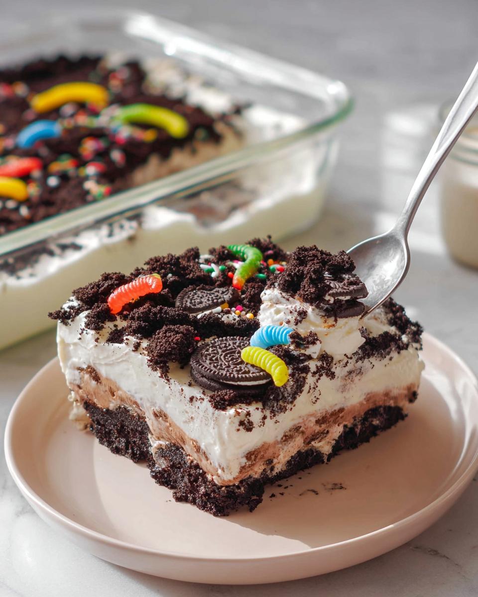 A spoonful is taken from a layered slice of Birthday Dirt Cake topped with gummy worms and Oreo crumbs.