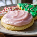 A close-up of a round sugar cookie topped with thick, swirled pink Simple Sugar Cookie Frosting, next to decorated holiday cookies.