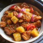 Close-up of saucy Slow Cooker Pineapple Chicken chunks with pineapple, red onion, and sesame seeds.