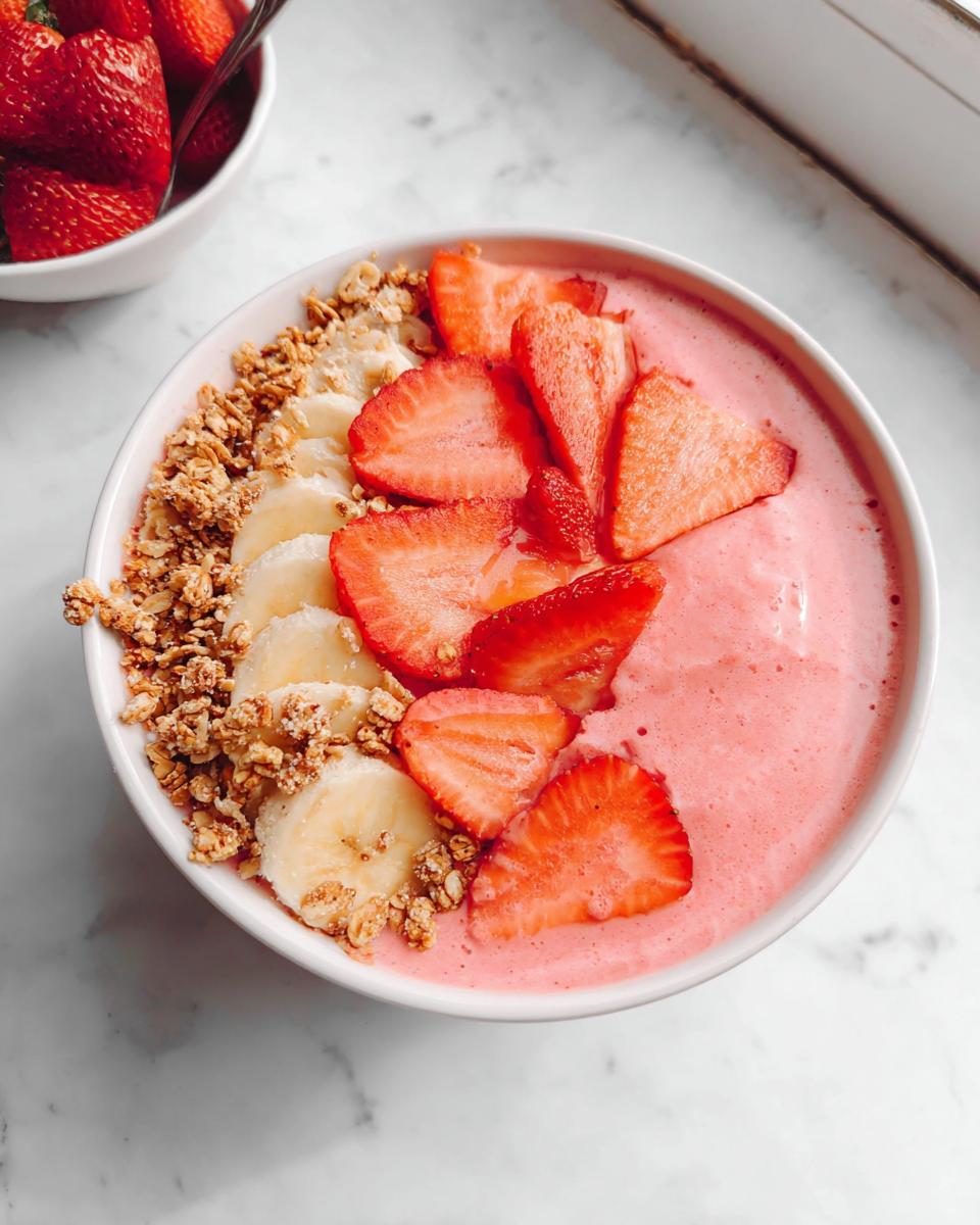 Overhead view of a vibrant pink Strawberry Banana Protein Smoothie Bowl topped with fresh strawberries, banana slices, and granola.