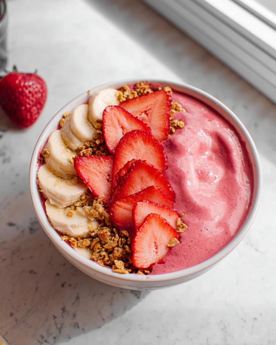 Close-up of a vibrant pink Strawberry Banana Protein Smoothie Bowl topped with sliced strawberries, banana coins, and granola.
