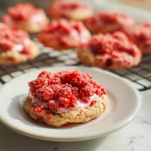 A close-up of one Strawberry Crumble Cookie topped with pink frosting and bright red crumble, sitting on a white plate.