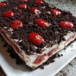A rectangular slice of Strawberry Dirt Cake featuring layers of cream, strawberries, and topped with chocolate cookie crumbs and whole strawberries.