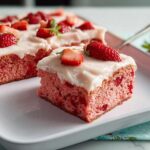 A close-up of a moist slice of pink Strawberry Sheet Cake topped with white frosting and fresh strawberry pieces.