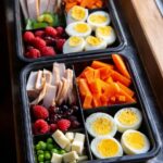 Two black compartmentalized containers filled with healthy Meal Prep Snack Boxes including sliced turkey, hard-boiled eggs, carrots, raspberries, and cheese.