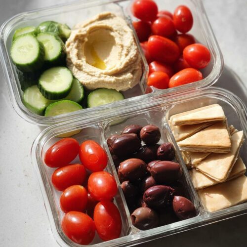 Two glass containers showing components of a Veggie & Hummus Lunch Box: hummus, cucumbers, tomatoes, olives, and pita bread.
